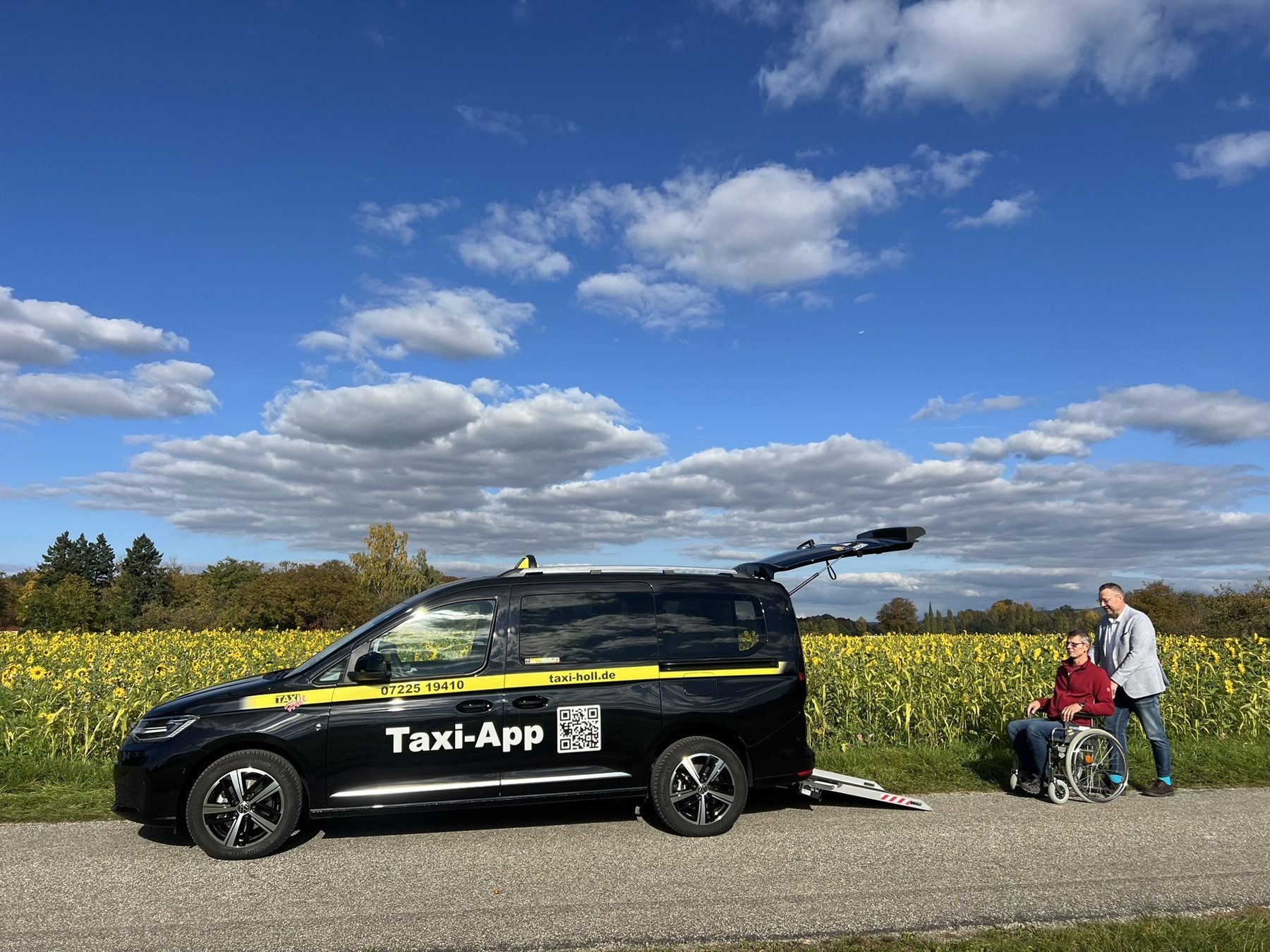 Schwarzes Taxi von Taxi-Holl mit ausgeklappter Heckrampe, daneben ein Fahrgast im Rollstuhl und Dirk Holl, dahinter ein Sonnenblumenfeld