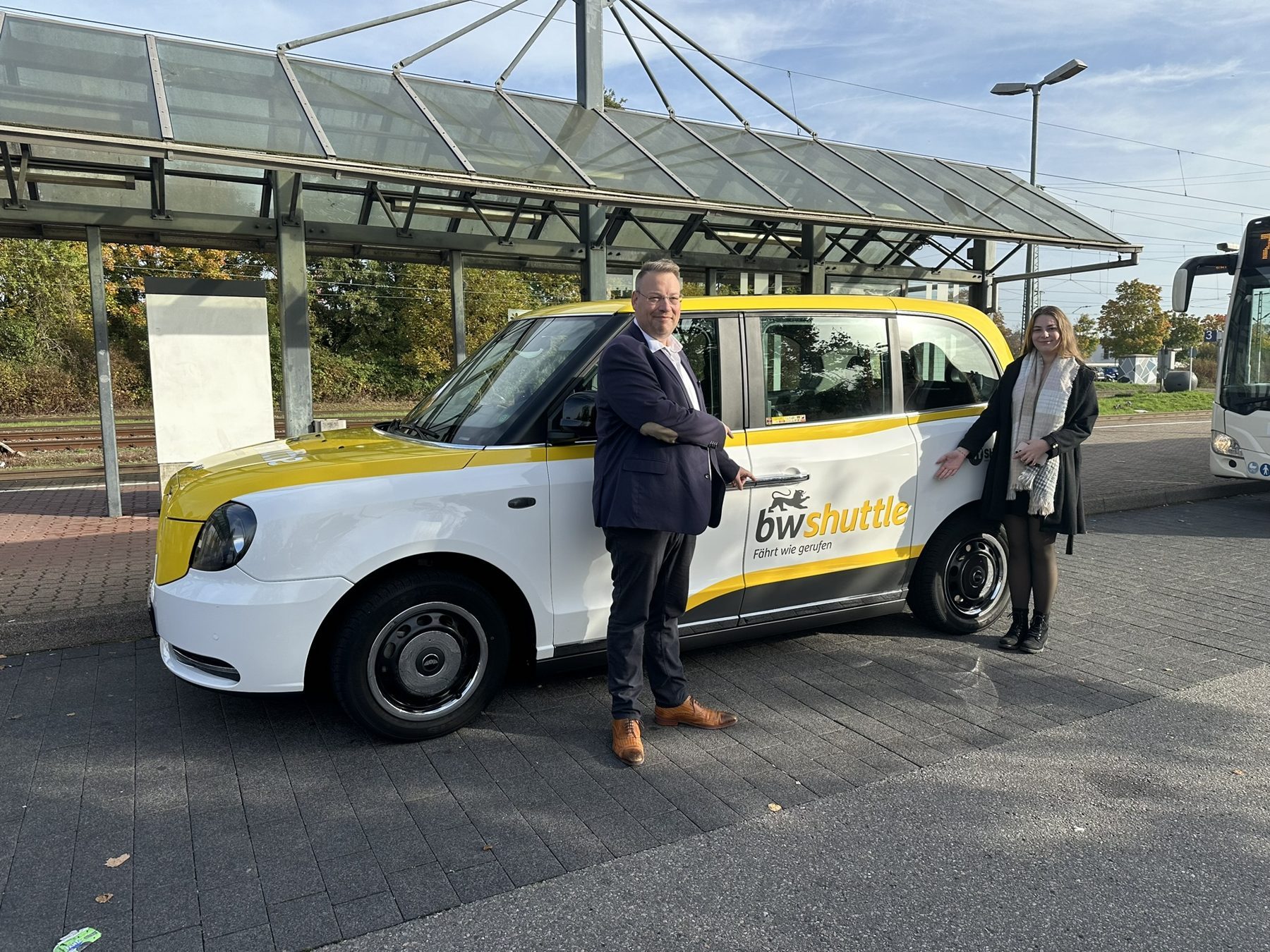 Dirk Holl und Romy Holl neben einem weiß-gelben London-Taxi mit der Aufschrift bwshuttle – Fährt wie gerufen an einer Bushaltestelle