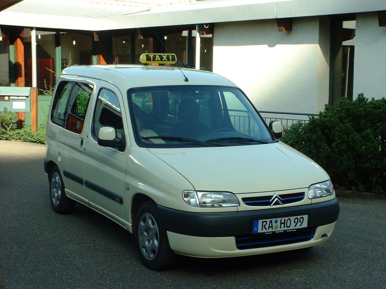 Hellelfenbeinfarbenes Taxi mit gelbem Dachschild, ein Hochdachkombi, vor einem Gebäude geparkt
