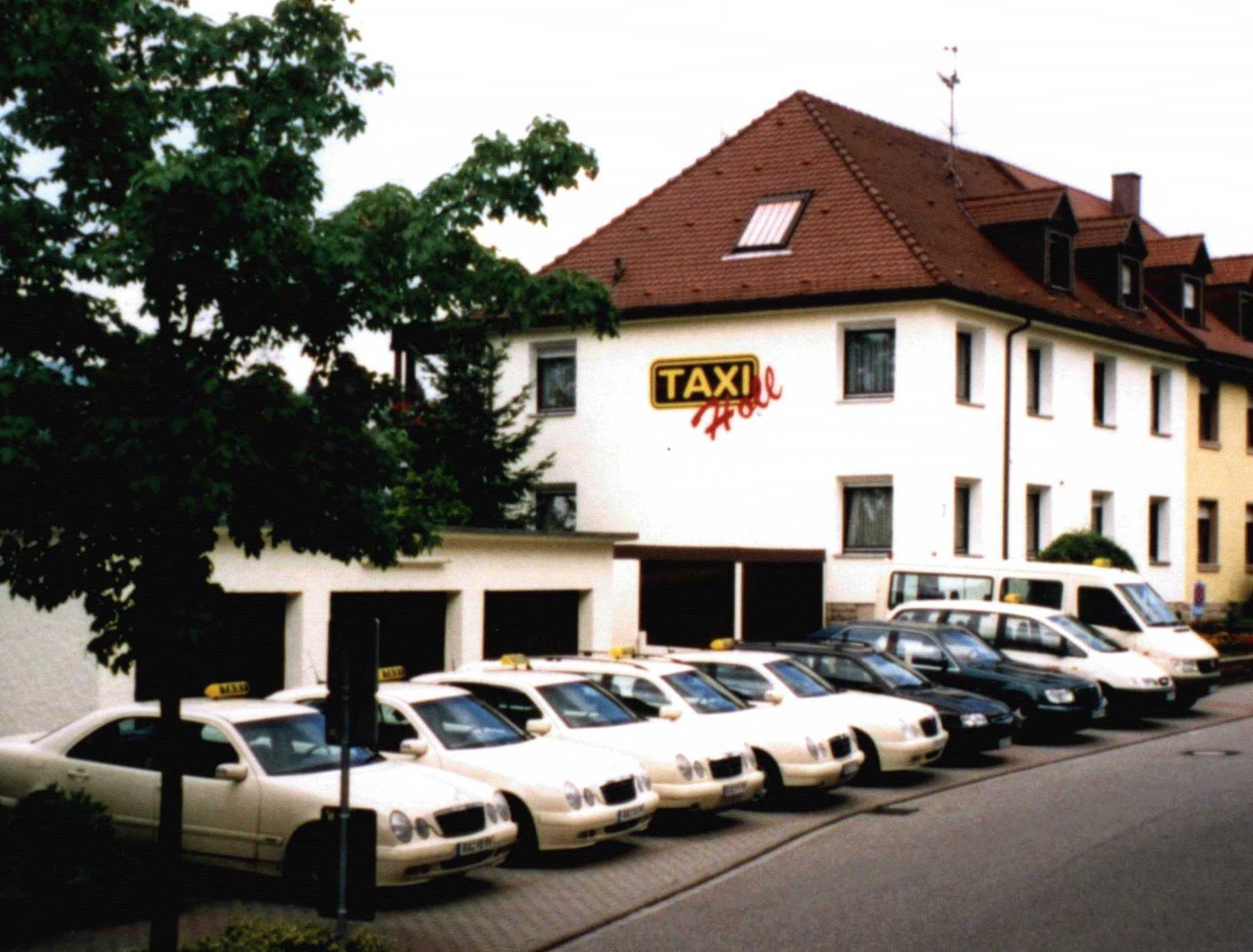 Weißes Gebäude mit Taxi-Holl-Schriftzug, davor eine Reihe elfenbeinfarbener Taxis und zwei Kleinbusse