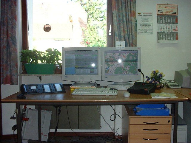 Büroarbeitsplatz mit zwei Röhrenmonitoren, links eine Auftragsliste, rechts eine Karte mit Fahrzeugpositionen, davor Telefonanlage und Funkgerät mit Mikrofon