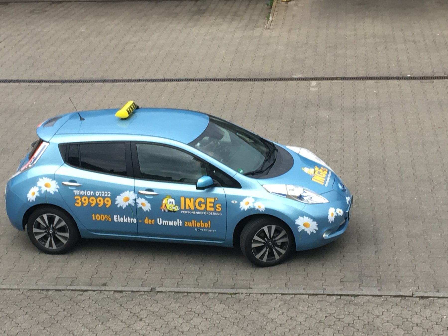 Hellblaues Elektroauto mit Taxischild, Inges-Logo, Gänseblümchen und der Aufschrift 100 % Elektro – der Umwelt zuliebe, von oben fotografiert