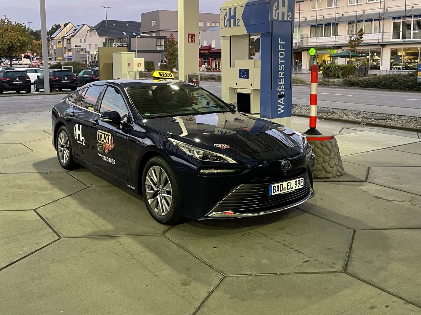 Dunkelblaues Taxi mit gelbem Dachschild, H2-Aufschrift und Taxi-Holl-Logo vor einer Zapfsäule mit der Aufschrift Wasserstoff