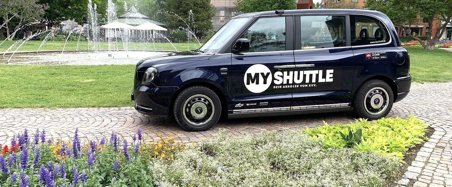 Dunkelblaues London-Taxi mit der Aufschrift MyShuttle – Dein Abholer vom KVV vor einem Park mit Springbrunnen