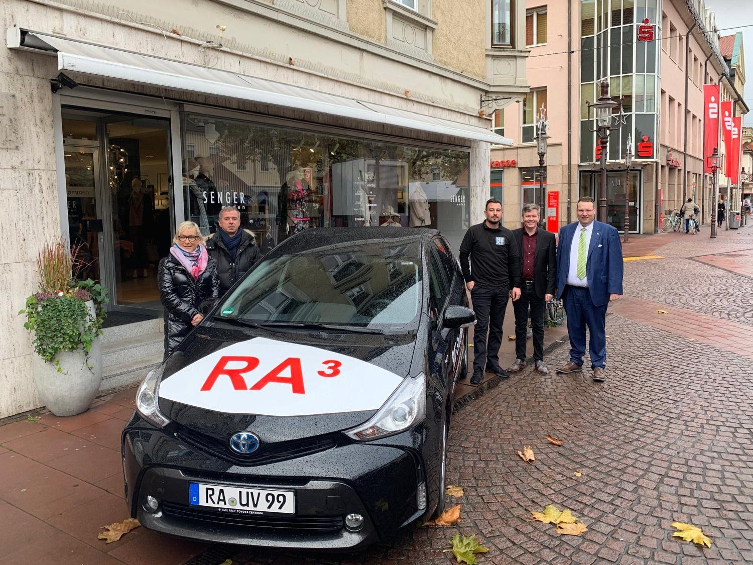 Das RA³-Shuttle, ein schwarzes Hybridfahrzeug mit RA³-Aufdruck auf der Motorhaube, vor einem Geschäft in der Rastatter Fußgängerzone, daneben fünf Projektbeteiligte