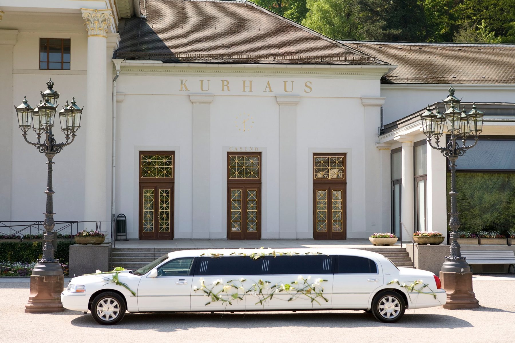 Weiße Stretchlimousine mit Hochzeitsblumenschmuck quer vor dem Kurhaus Baden-Baden mit dem Schriftzug Kurhaus und zwei Kandelabern