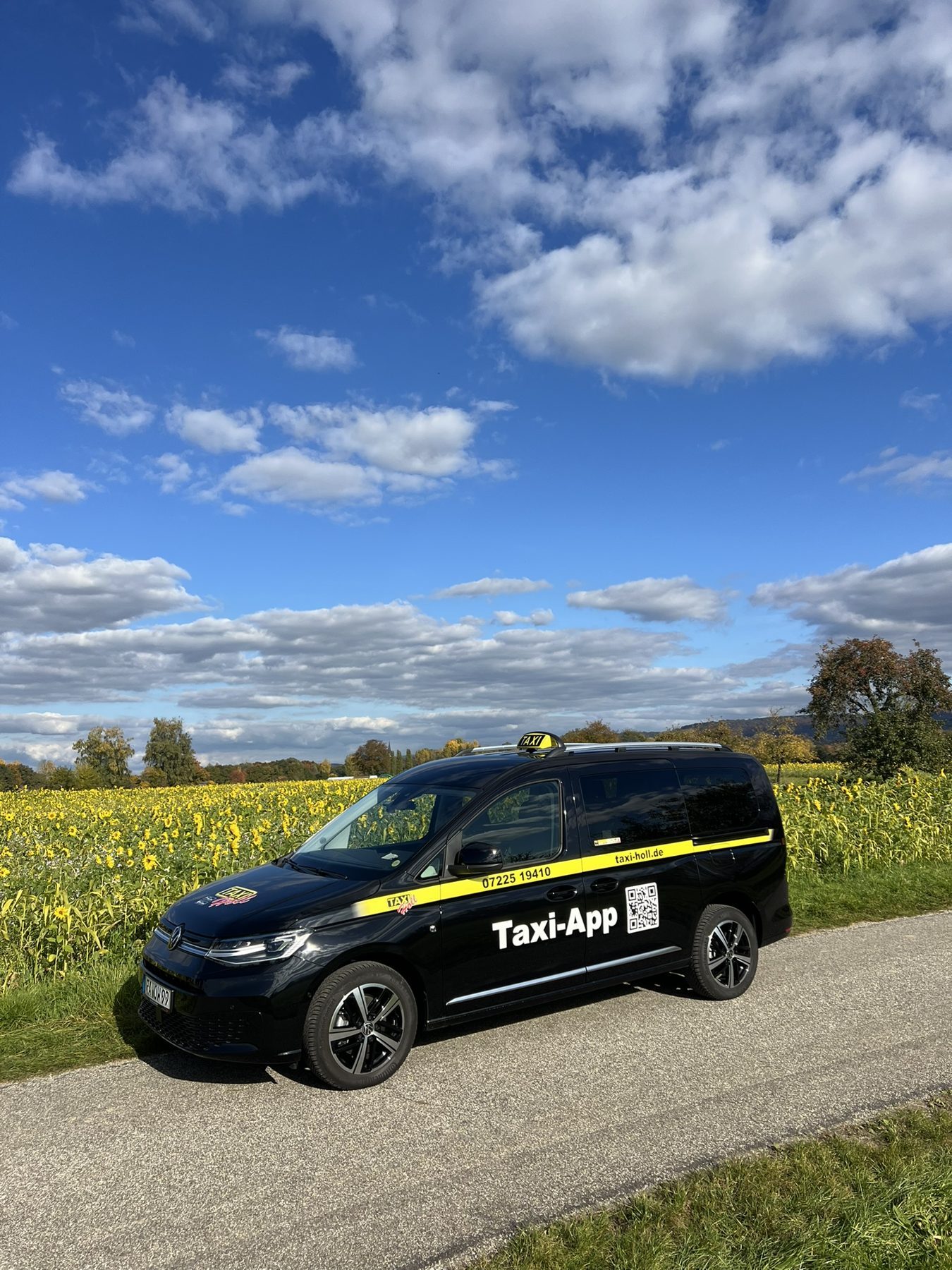 Schwarzes Taxi von Taxi-Holl mit gelbem Streifen vor einem Sonnenblumenfeld unter blauem Himmel