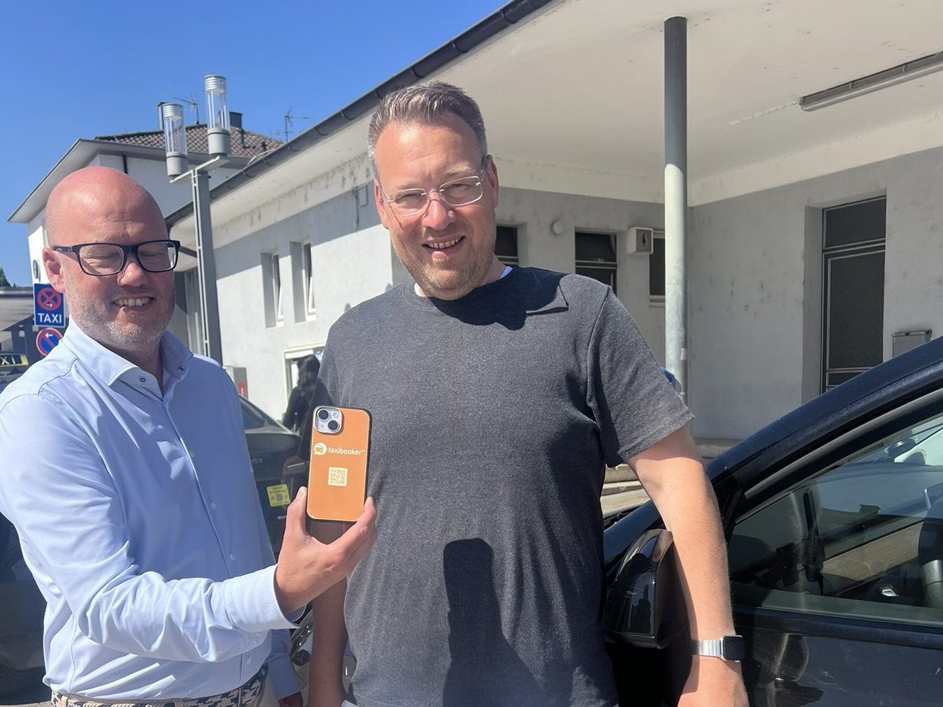Arnoud de Niet zeigt ein Handy mit Taxibooker-Hülle und QR-Code, daneben Dirk Holl an einem Taxi von Taxi-Holl