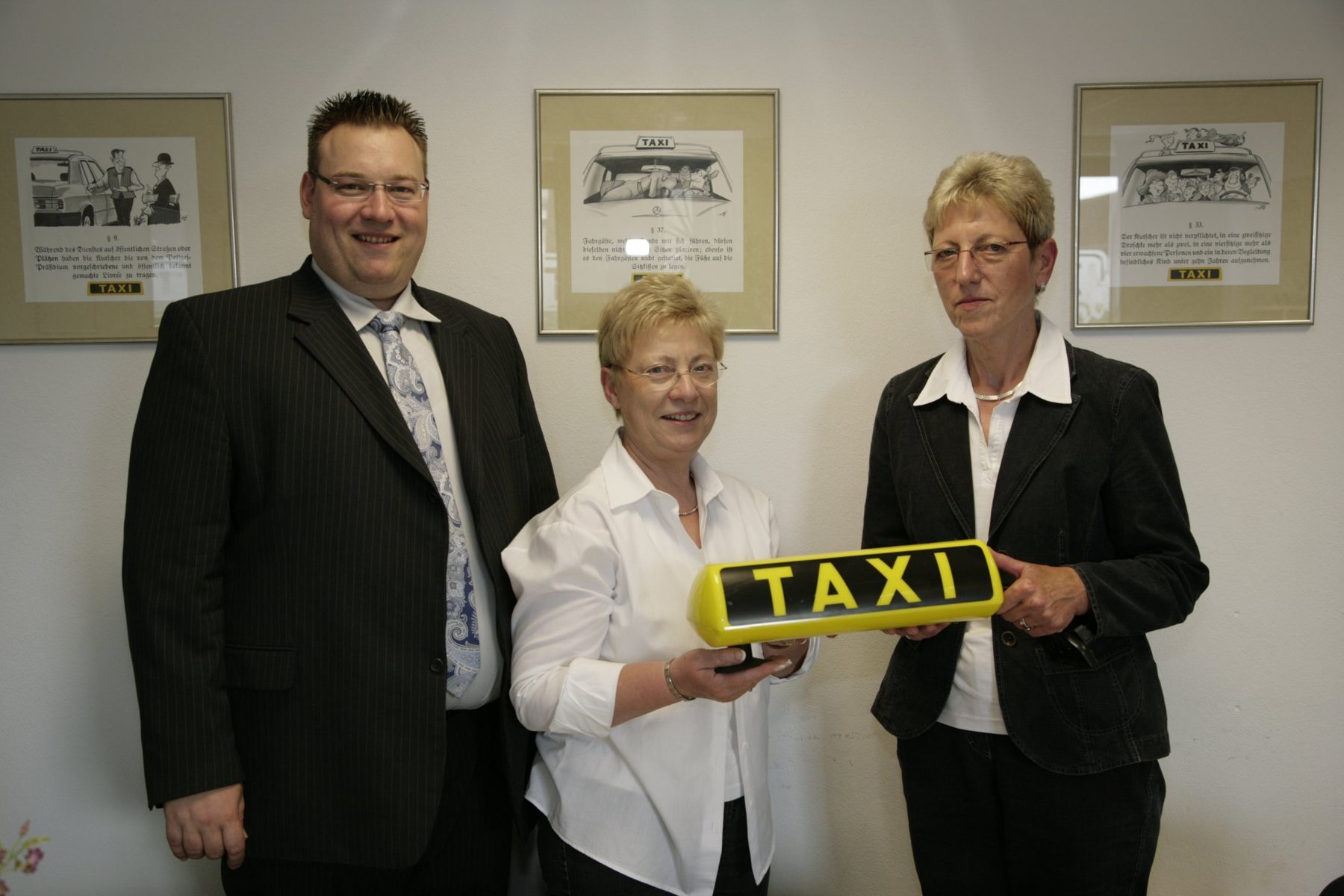 Dirk Holl, Edeltraud Holl und Frau Schuon halten gemeinsam ein gelbes Taxischild, an der Wand gerahmte Taxi-Karikaturen