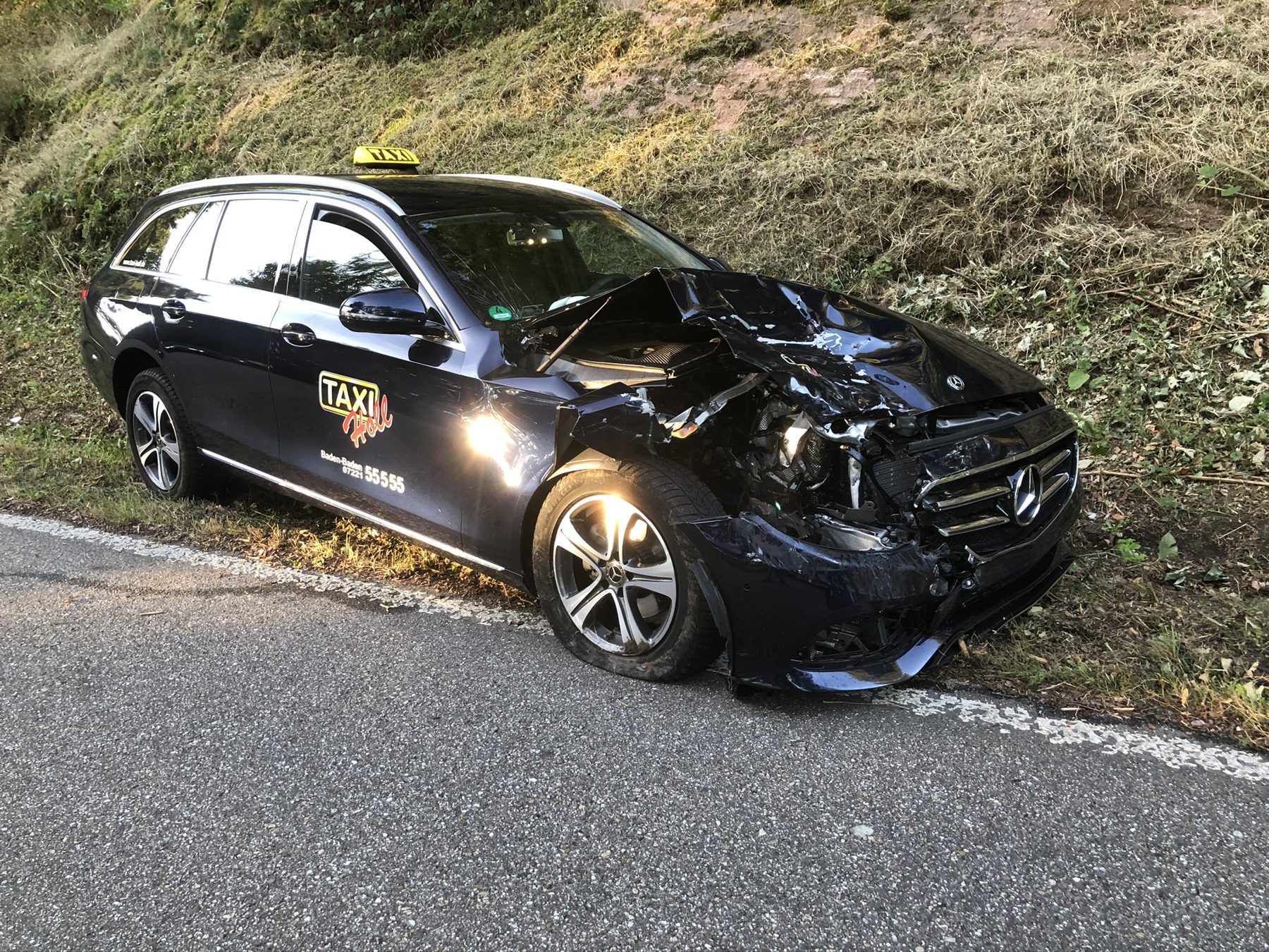Schwarzes Taxi von Taxi-Holl mit völlig zerstörter Front am Straßenrand vor einer Böschung